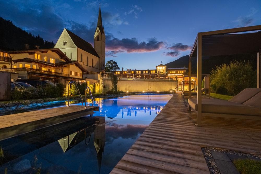 Hotelpool mit Holzdeck bei Abendstimmung neben Kirche