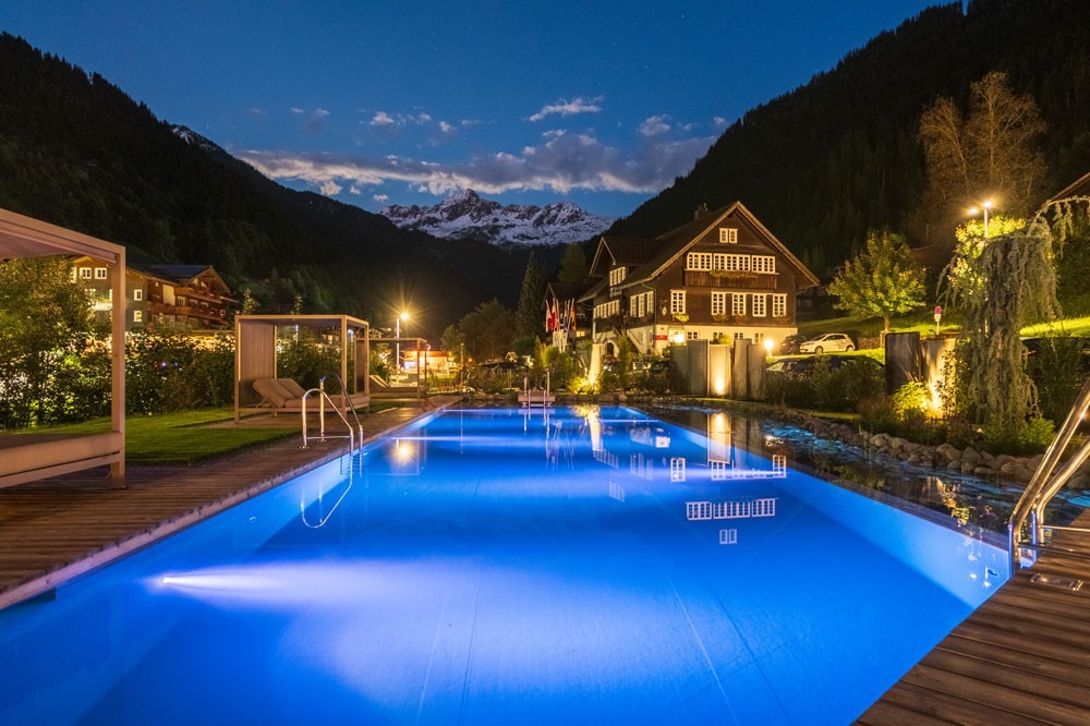 Beleuchteter Hotelpool bei Nacht vor Bergdorf und Alpenkulisse