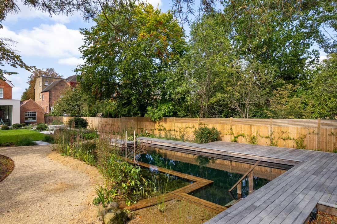 Naturpool mit Holzsteg vor modernem Gartenhaus im grünen Garten