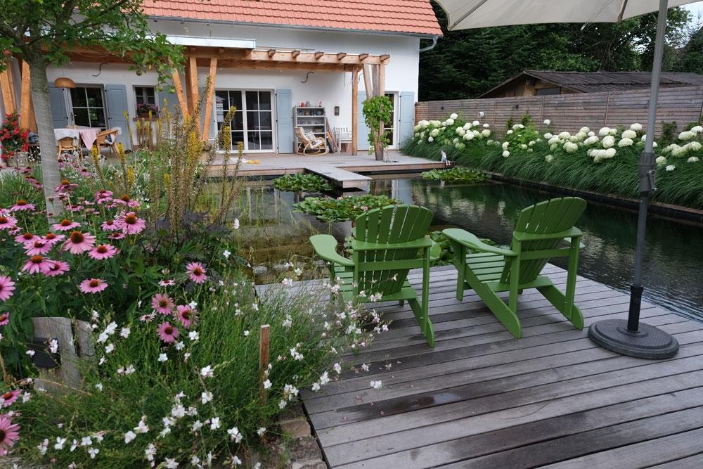 Naturpool mit Holzdeck gruenen Sesseln und bluehendem Garten