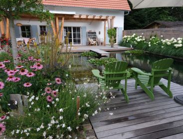 Naturpool mit Holzdeck gruenen Sesseln und bluehendem Garten