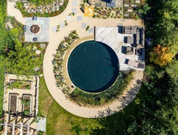 Luftaufnahme eines runden Schwimmteichs mit vielfaeltiger Bepflanzung und Holzterrasse